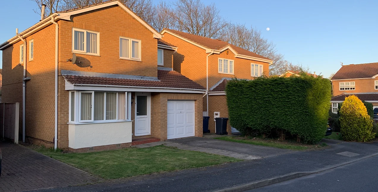 Cloverlands House, a detached brick house in a suburban neighbourhood. Trees can be seen behind the house, with the moon rising behind them in a dark blue evening sky.