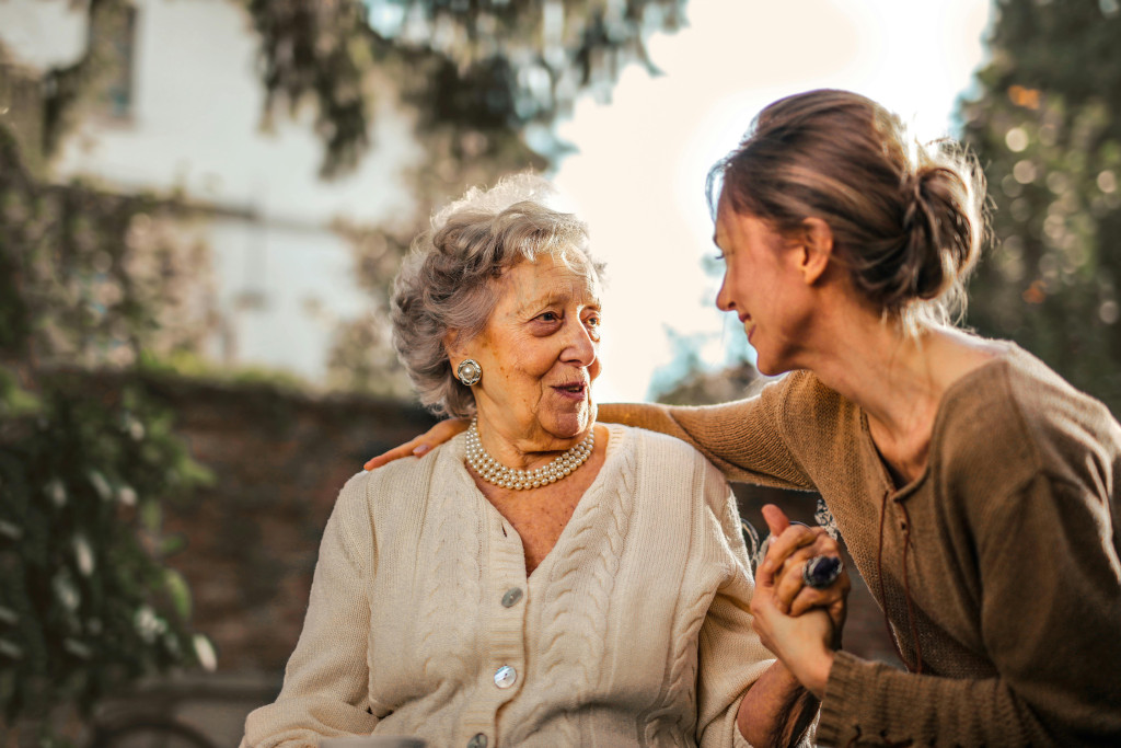 A woman comforting and caring for an older woman, putting an arm round her shoulders. The background is soft focus but shows they are outside, perhaps in a garden. Photo by Andrea Piacquadio