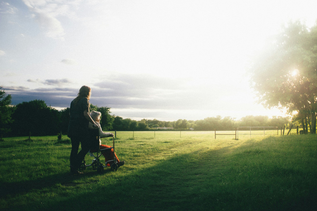 A man pushing an older woman in a wheelchair across a grass area, out of the long shadows and into the sunlight. Photo by Dominik Lange.